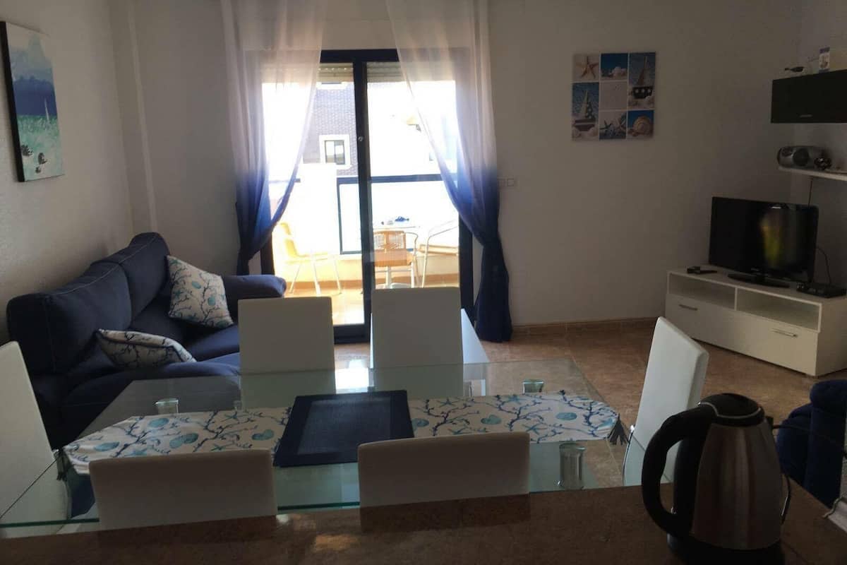 Una sala de estar con sofá, mesa de centro y televisor. Tiene un balcón con vistas al exterior.
