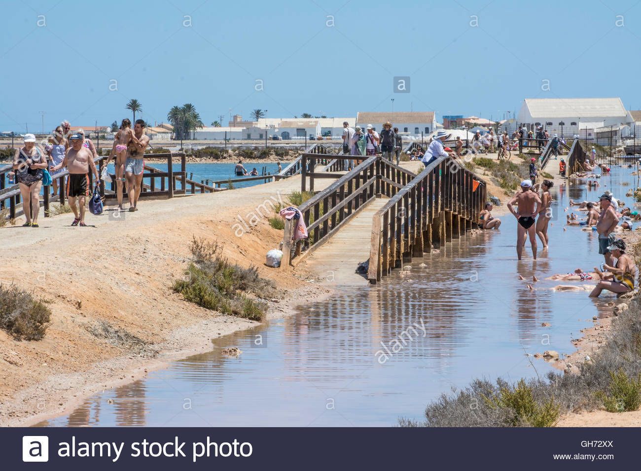 Escena de playa con un puente de madera, gente caminando y una zona de piscina.