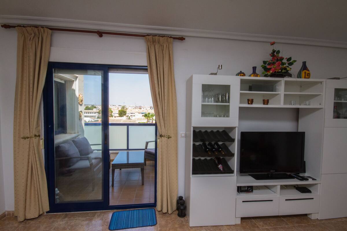 Un salón con televisor, estanterías y vistas al exterior.