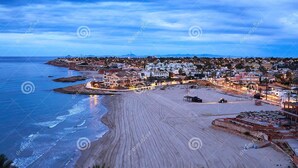 Une station balnéaire dotée d’une vaste plage de sable, d’un espace piscine avec des chaises longues, et offrant une vue sur l’océan et la ville en arrière-plan.