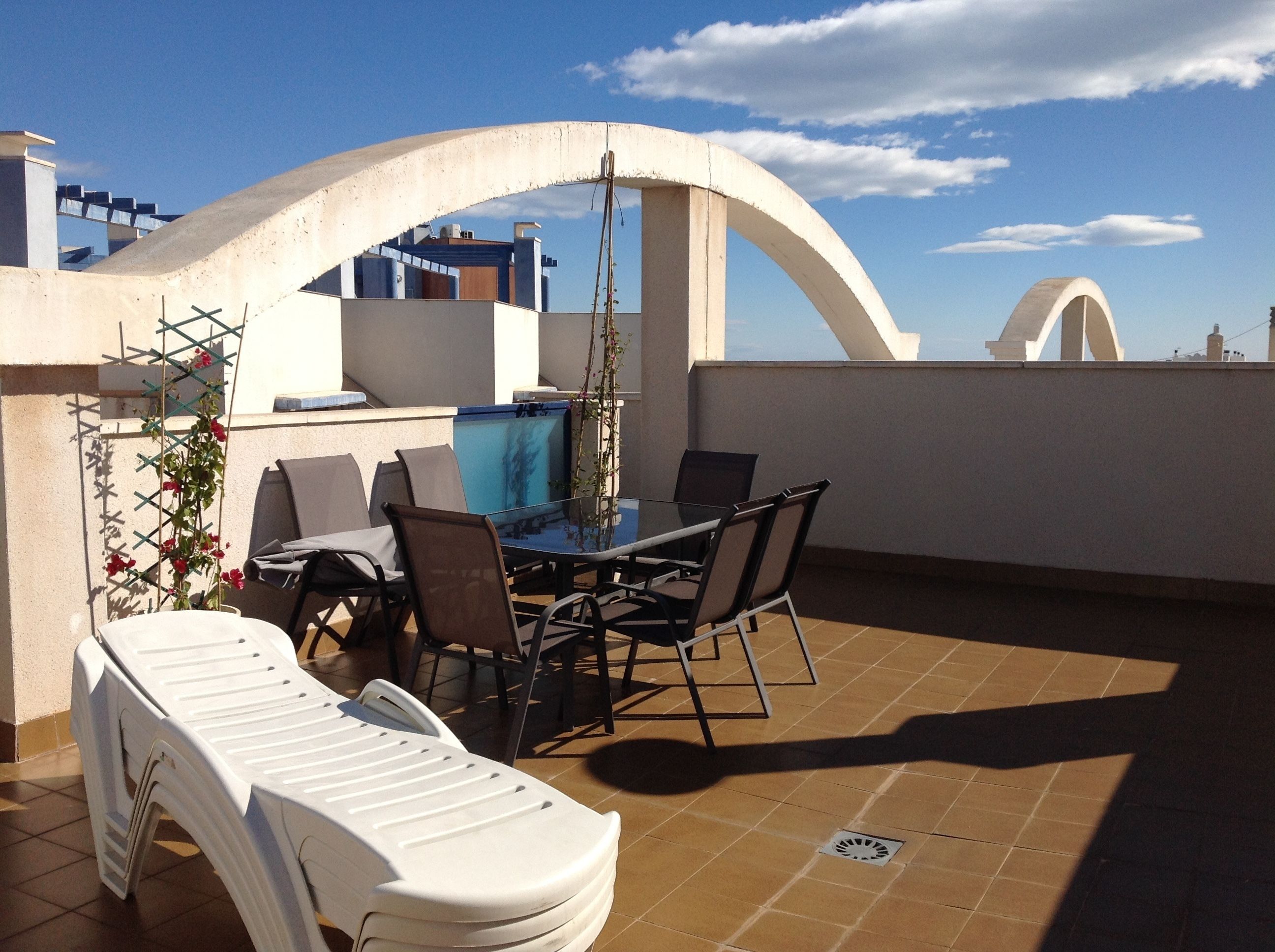 A rooftop terrace with a table and chairs, a white plastic chair, and a view of the cityscape.