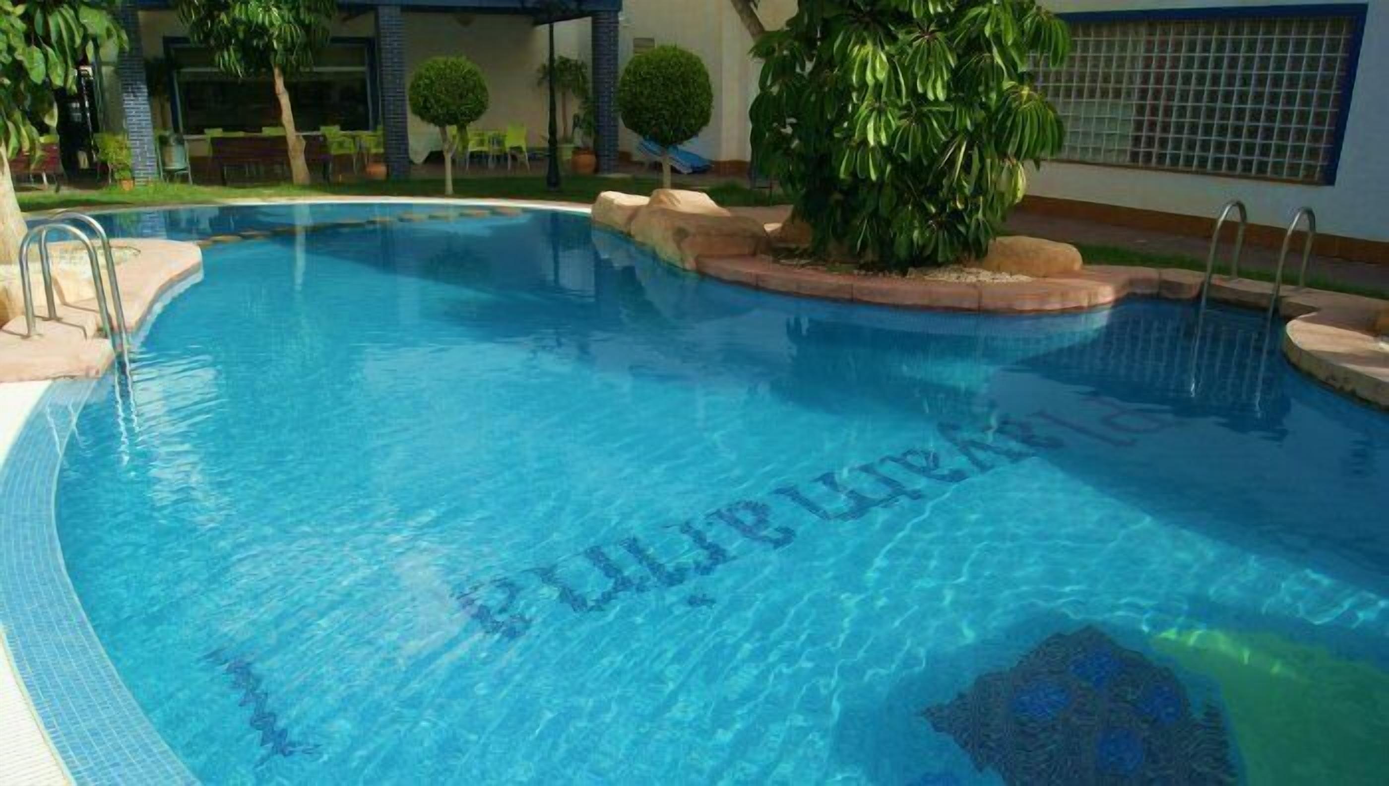 Una piscina azul cristalina con borde de azulejos, rodeada de jardines con árboles pequeños y un edificio al fondo.
