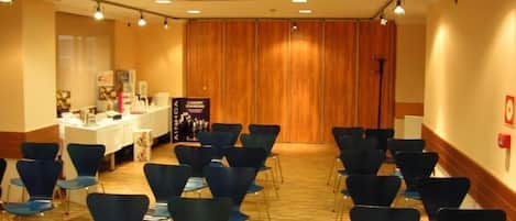 Un salón de conferencias con filas de sillas azules, una mesa blanca con un banner y paneles de madera al fondo.