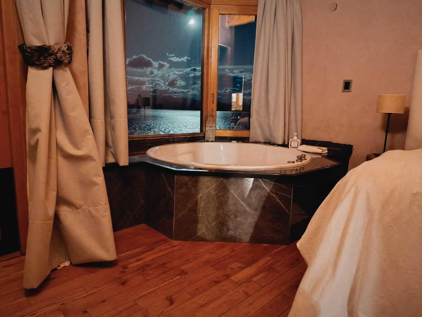 Une salle de bain avec une grande baignoire, un plancher en bois et une fenêtre avec des rideaux.
