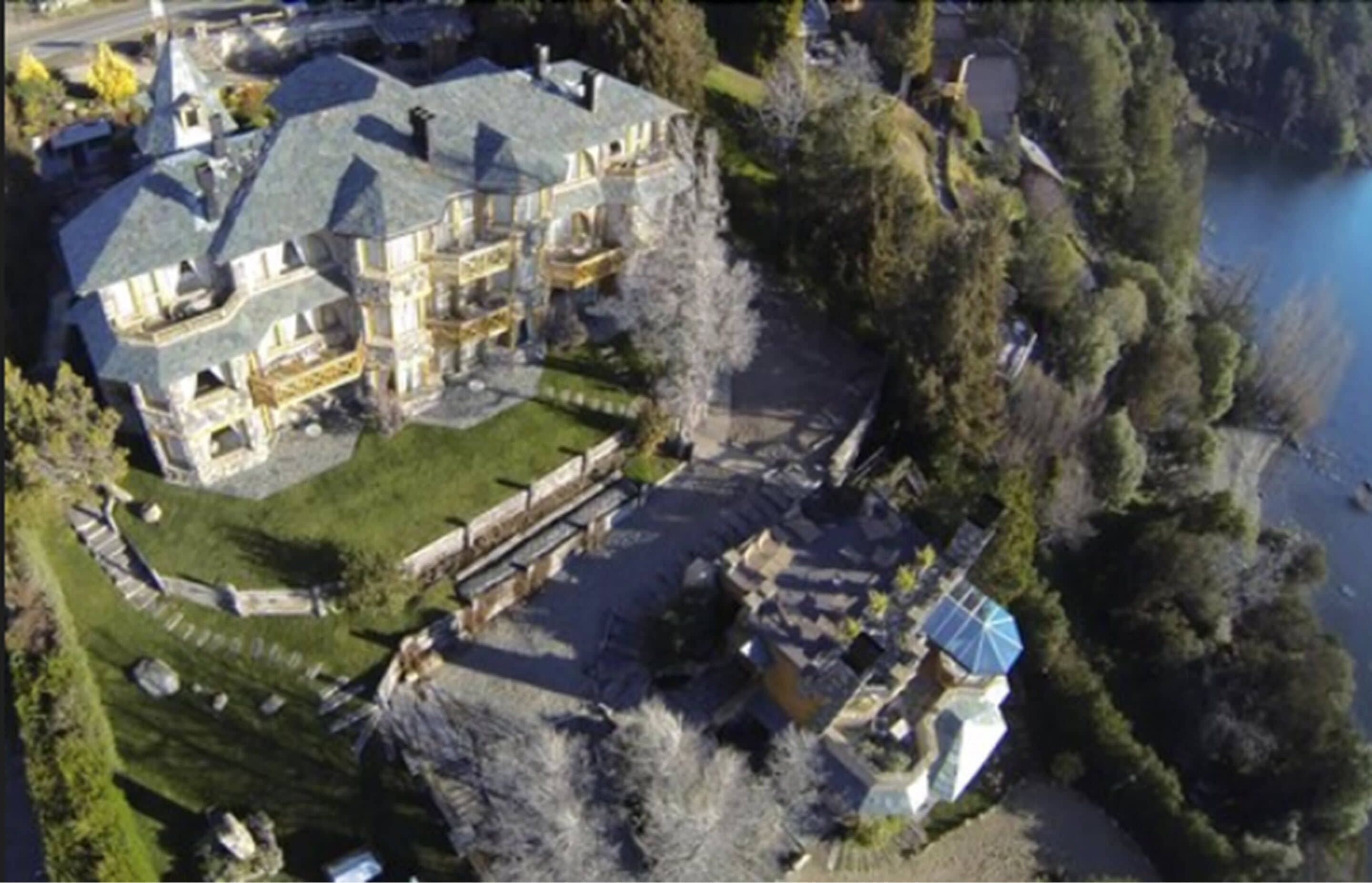 An aerial view of a large, multi-story house with a spacious lawn, a swimming pool, and a dock extending into a body of water.