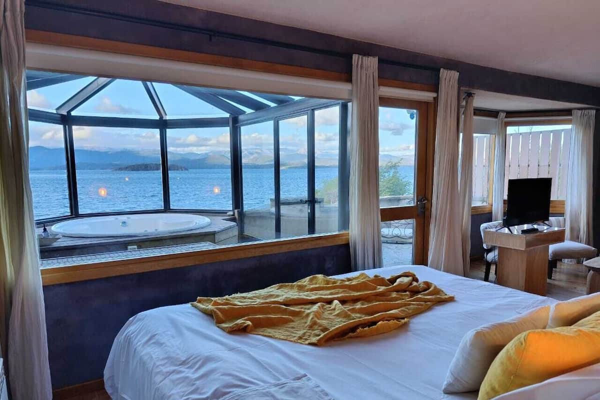 A bedroom with a large bed, a desk, and a view of a body of water through large windows.