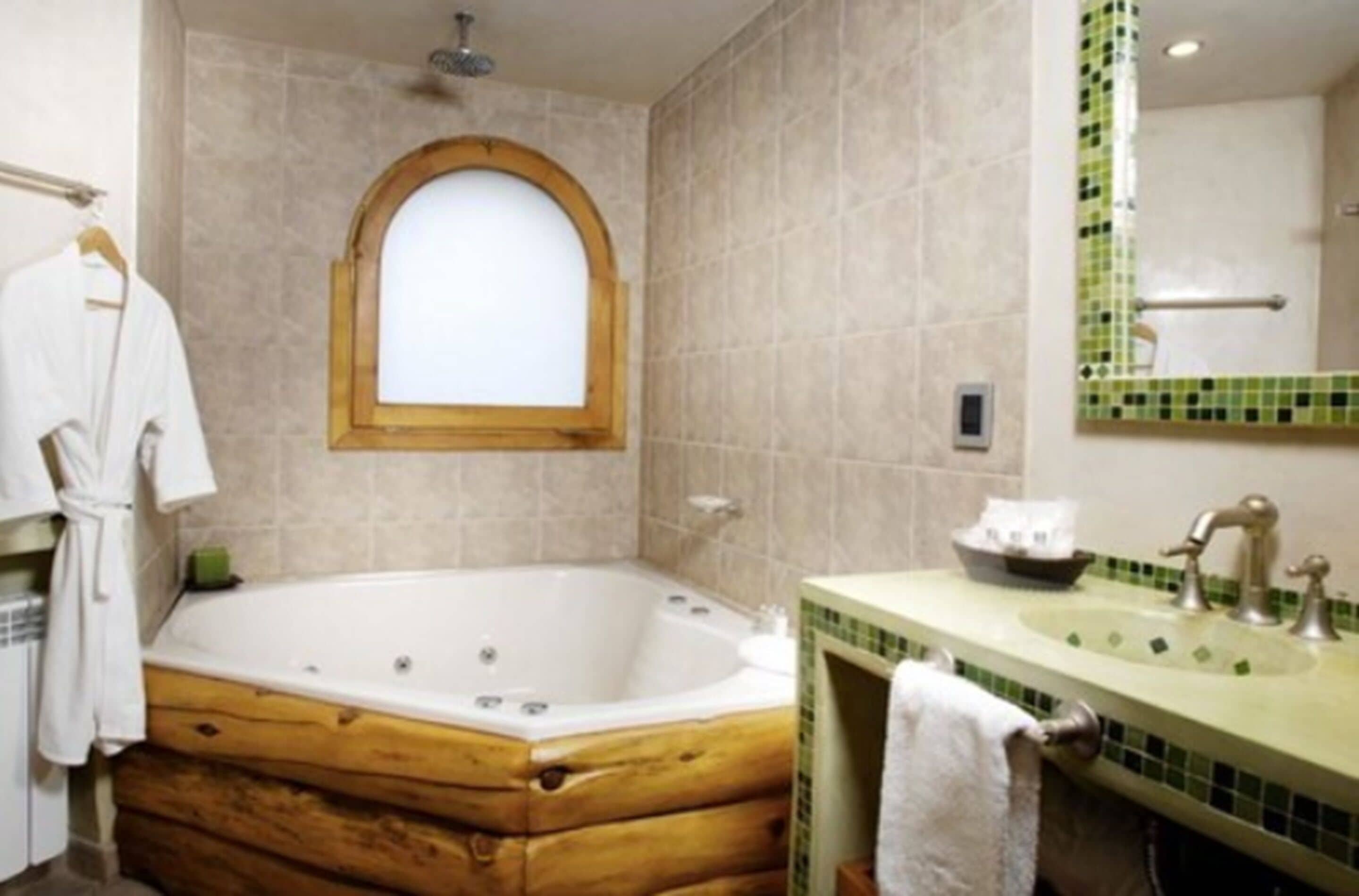 A bathroom with a wooden bathtub, a sink with a green-tiled countertop, and a mirror with a green border.