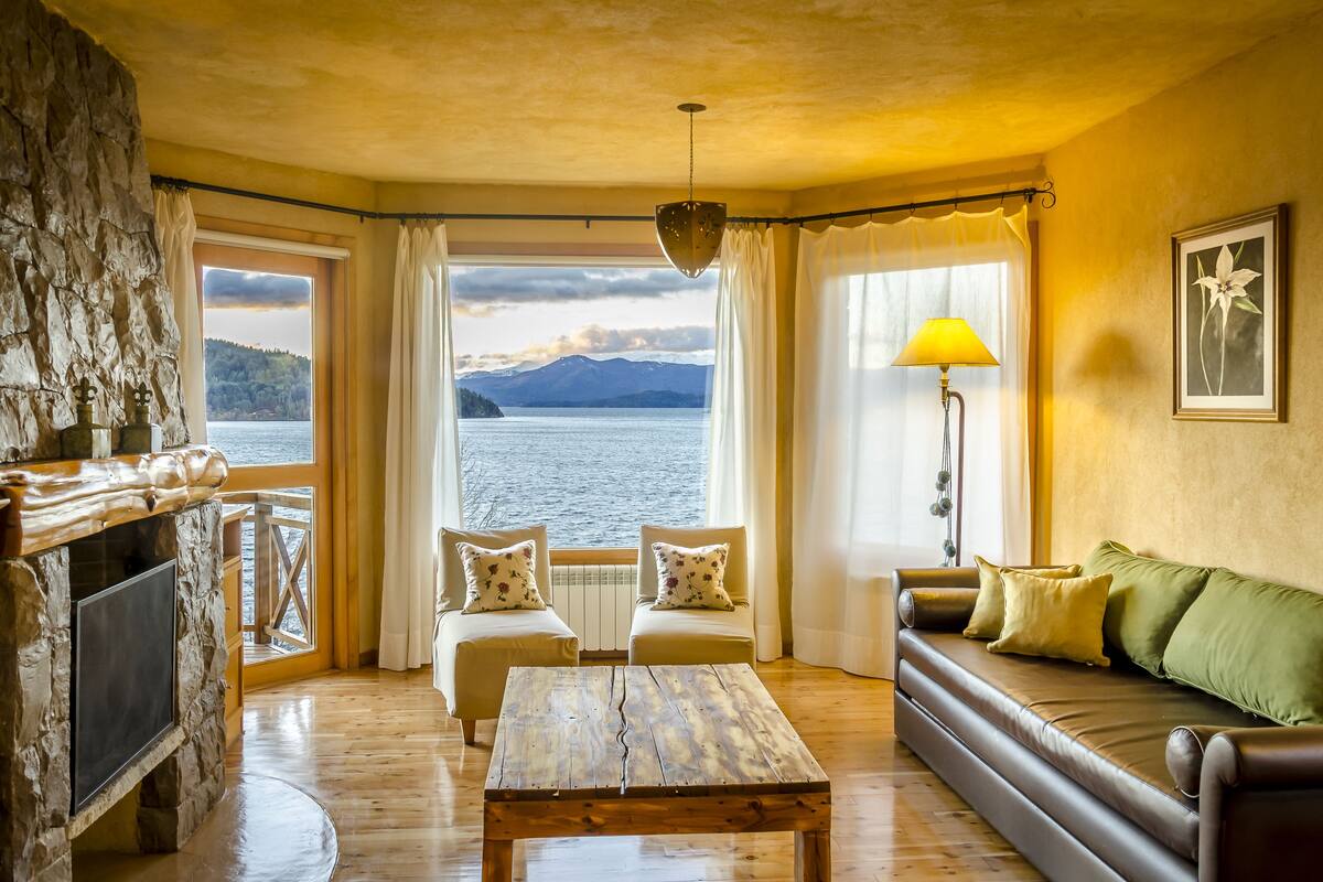 A cozy room with a stone fireplace, a wooden table, a sofa with cushions, and a view of a lake and mountains.