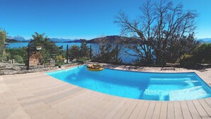 Piscina com deck de madeira, rodeada por árvores e vista para montanhas e um lago.