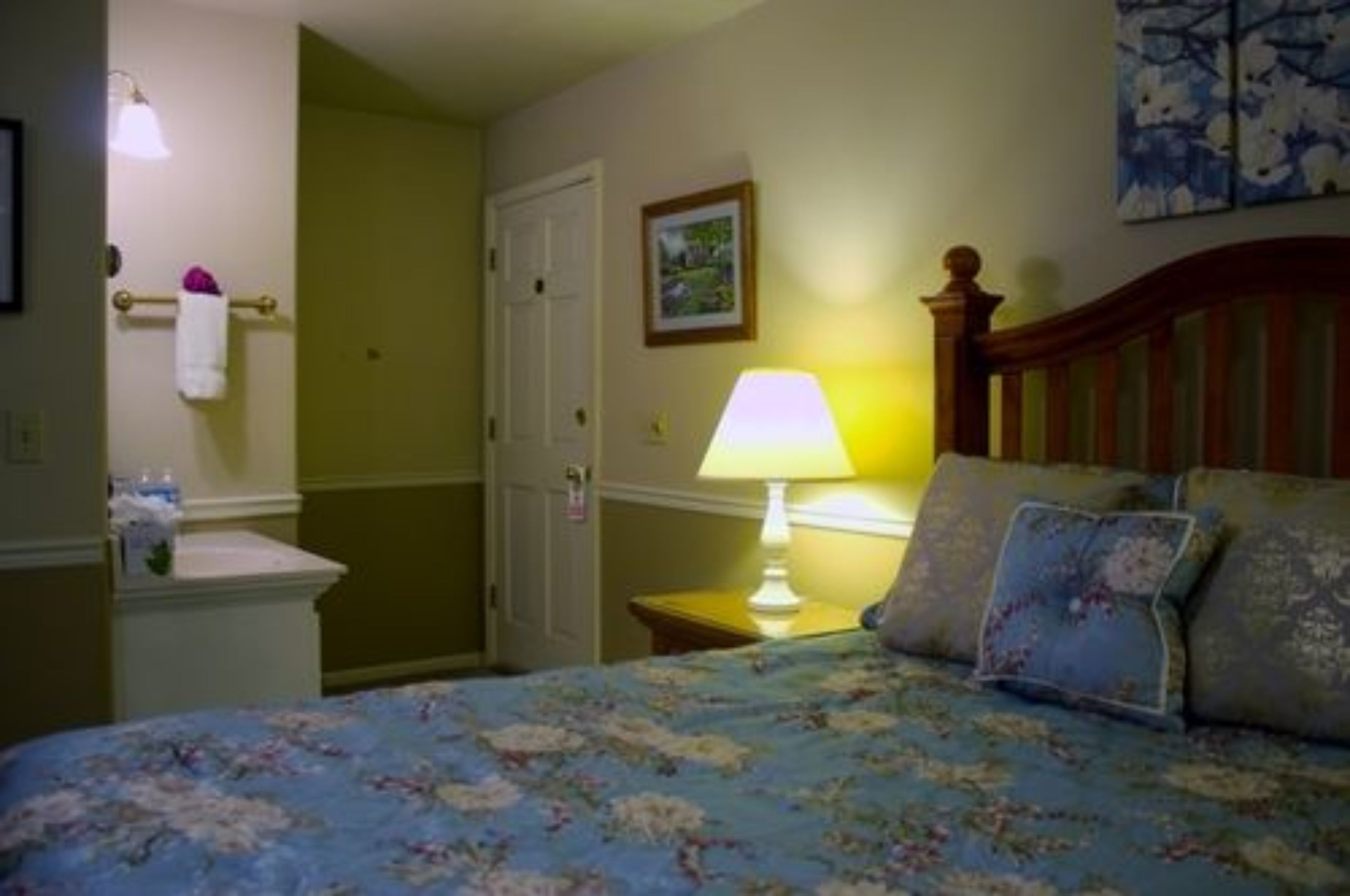 A bedroom with a bed, a nightstand, a lamp, a wall-mounted towel rack, and a door.