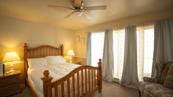 A bedroom with a wooden bed, a ceiling fan, two bedside lamps, a nightstand, a chair, and a window with curtains.