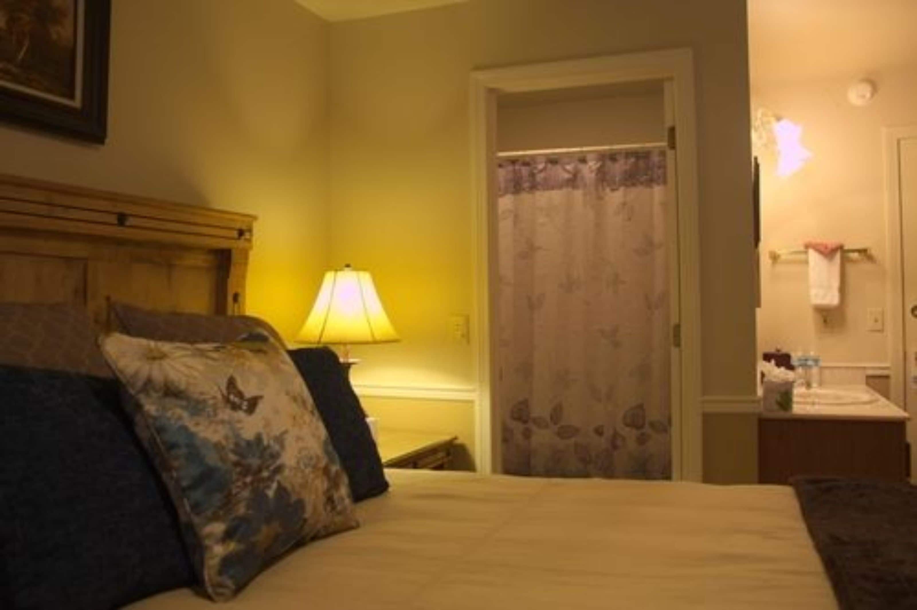 A hotel room with a bed, a nightstand with a lamp, a door with a floral curtain, and a small table with a towel and toiletries.