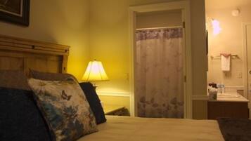A hotel room with a bed, a nightstand with a lamp, a door with a floral curtain, and a small table with a towel and toiletries.