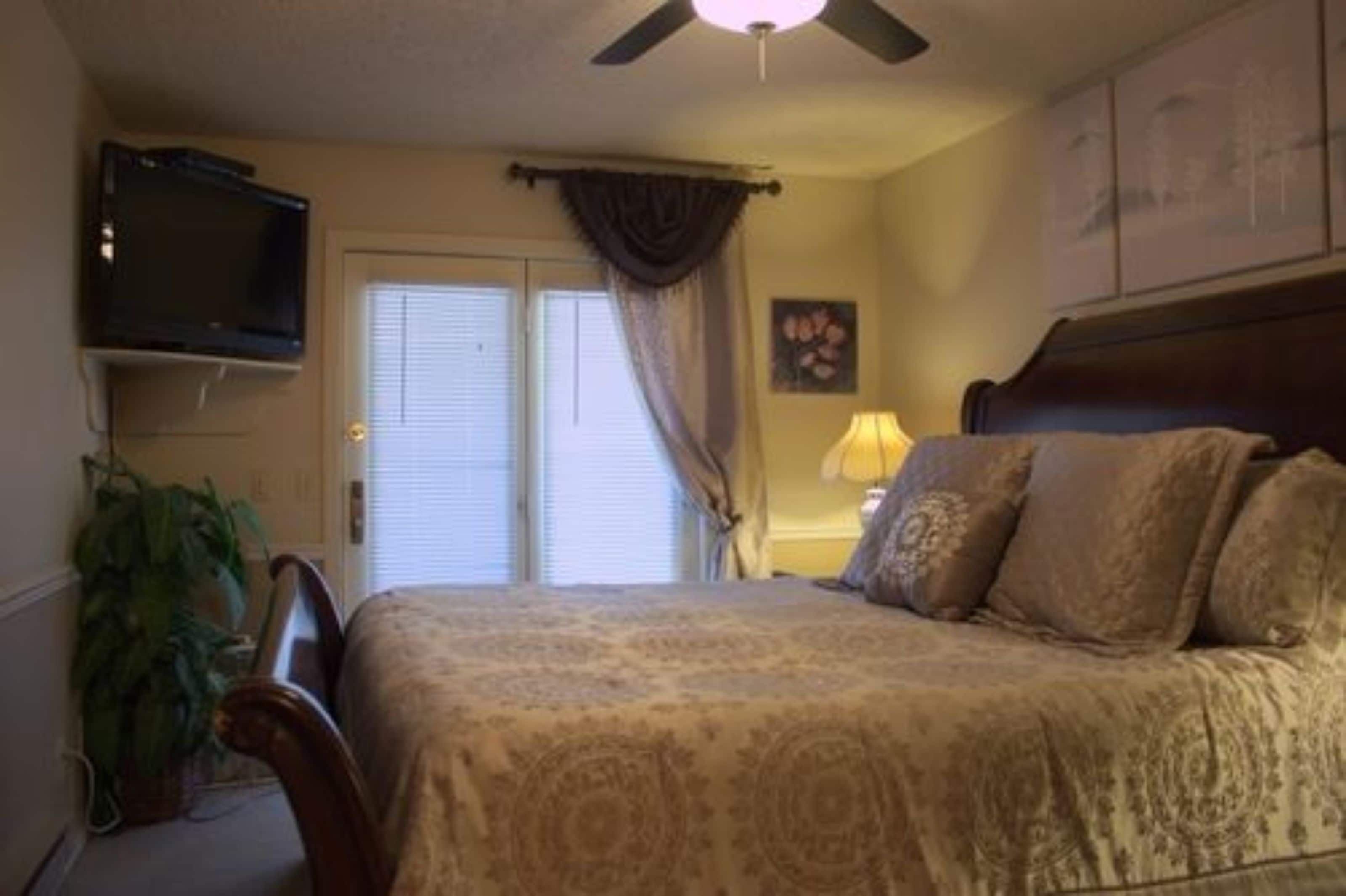 A bedroom with a bed, a television mounted on the wall, a ceiling fan, and a window with curtains.