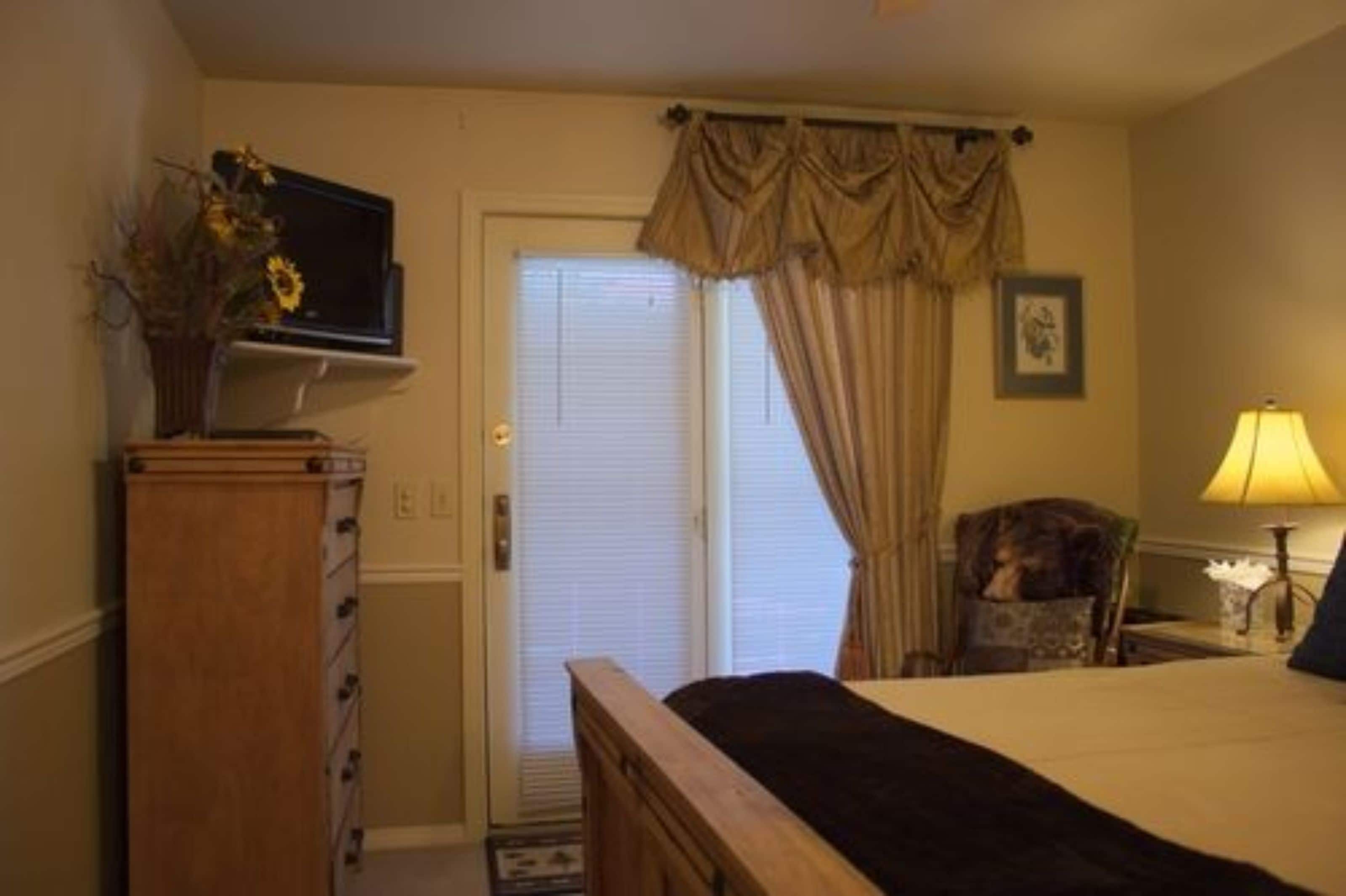 Une chambre à coucher avec un lit, un téléviseur posé sur un meuble, une chaise, une lampe et une porte avec des rideaux.