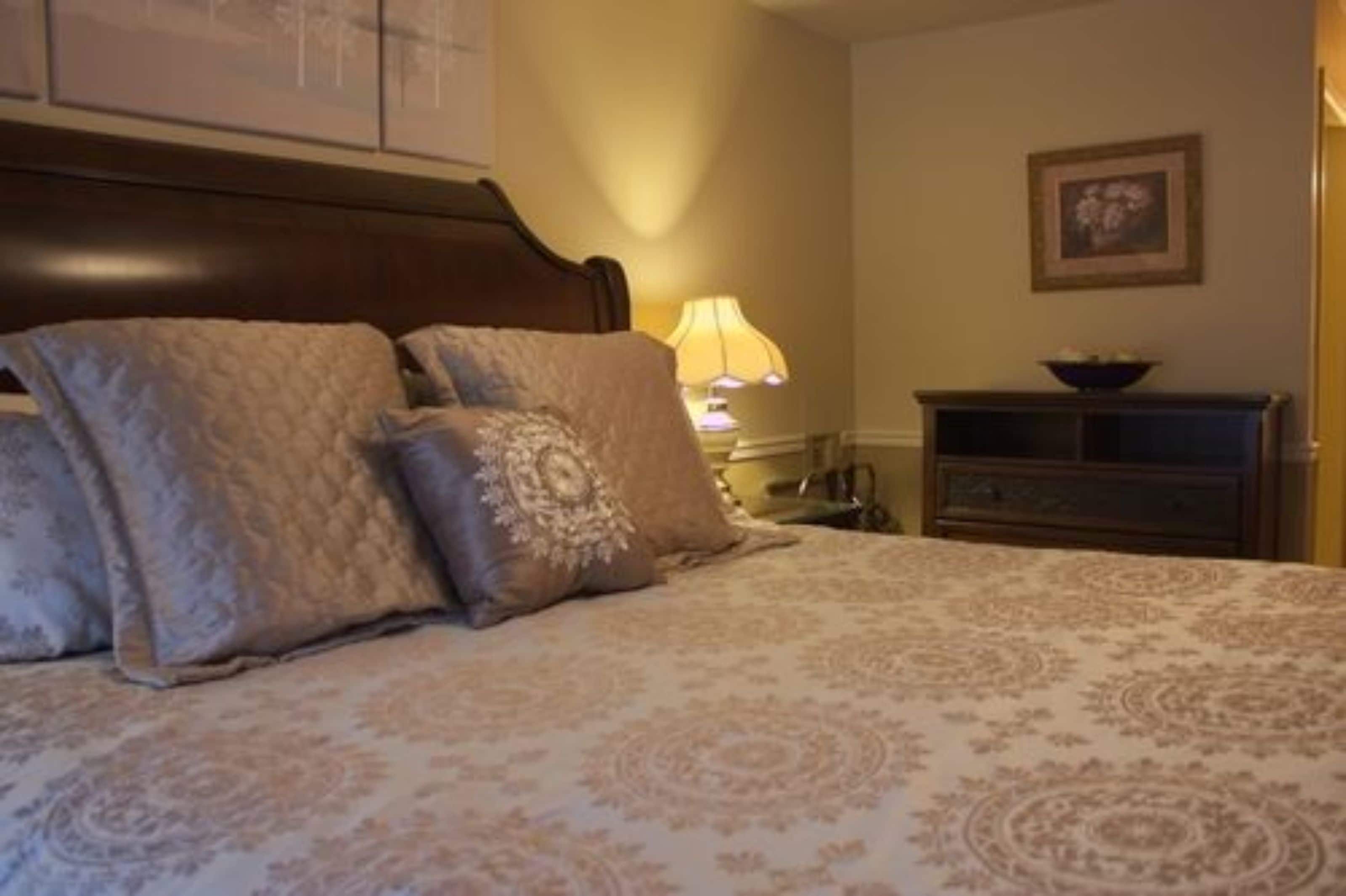 A neatly made bed with a patterned bedspread and two pillows, a bedside lamp, and a small wooden cabinet against a wall with a framed picture.