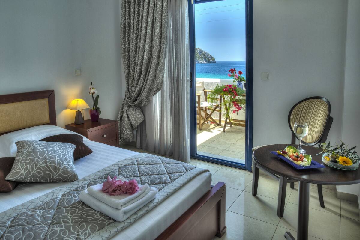 Une chambre d’hôtel avec un lit, une table de chevet avec une lampe, une chaise et une table sur laquelle se trouvent un panier de fruits et un verre d’eau.