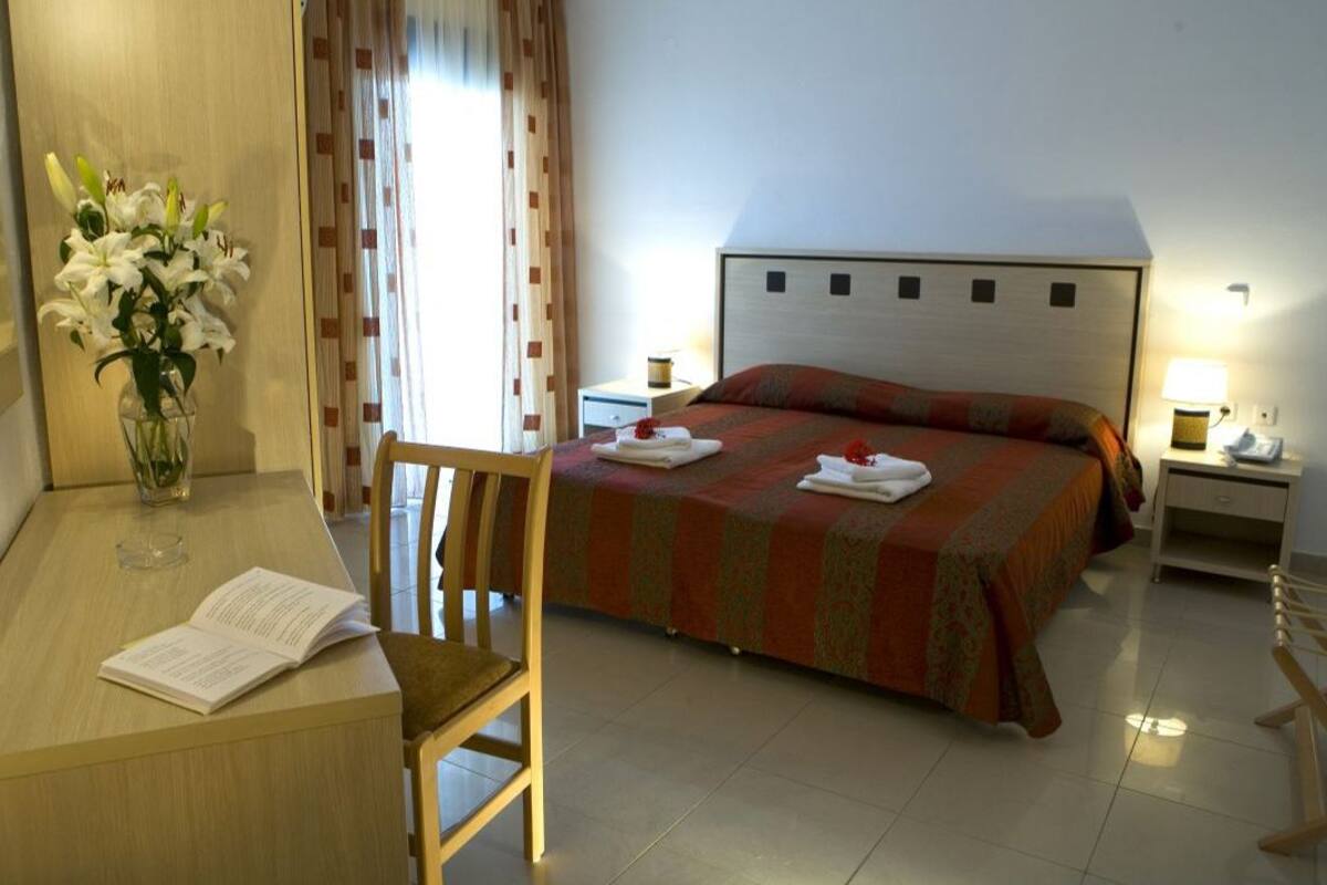 Une chambre d’hôtel comprenant un lit, un bureau avec une chaise, un vase de fleurs et une table de chevet avec une lampe.