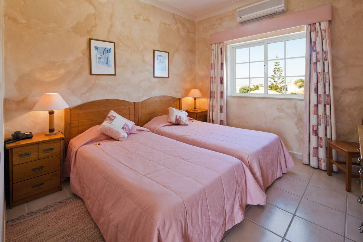Une chambre avec deux lits, chacun doté d’une table de chevet et d’une lampe, d’une commode en bois, d’une chaise et d’une fenêtre avec des rideaux.