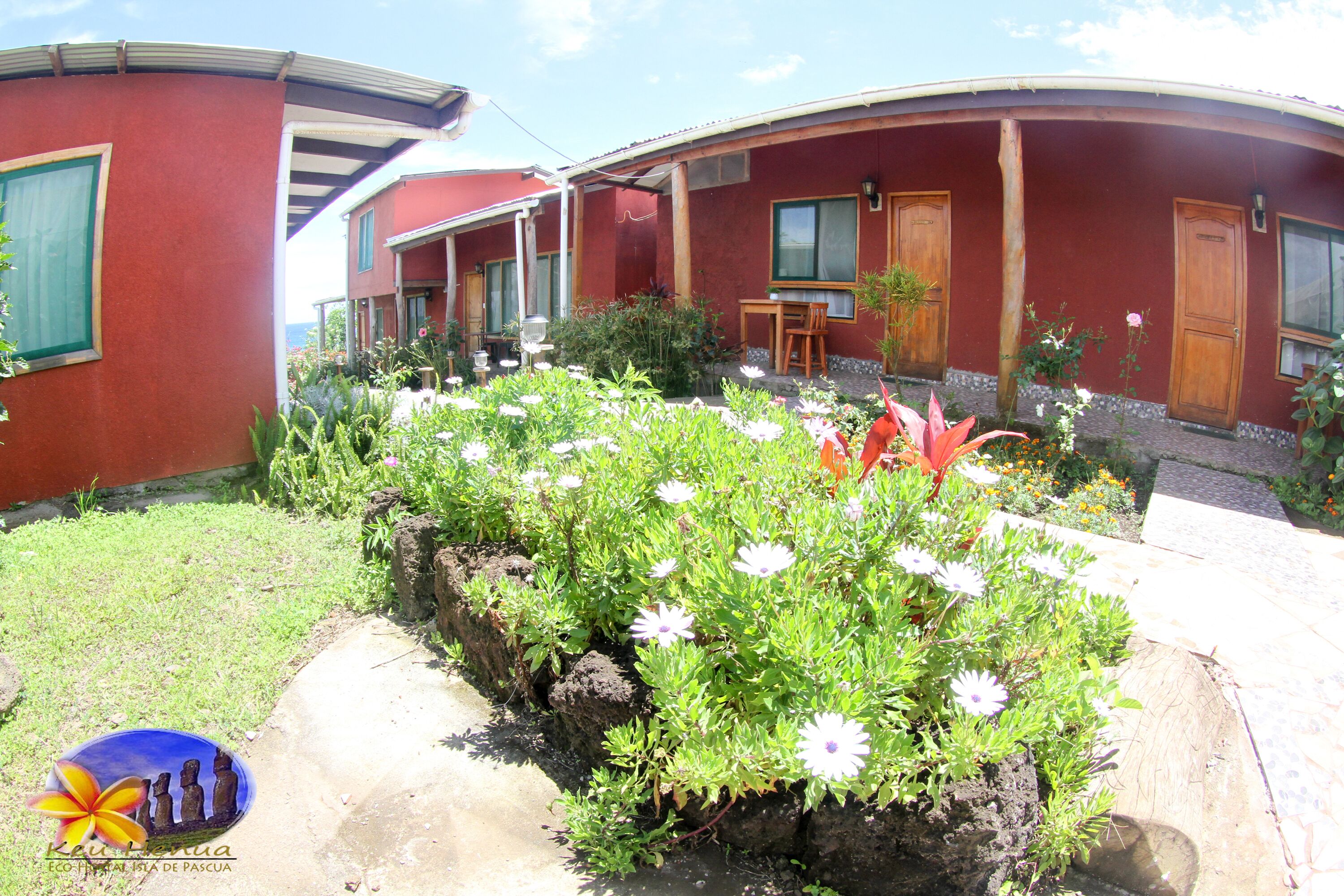 A red building with a covered patio area, a small garden with white flowers, and a logo in the bottom left corner.