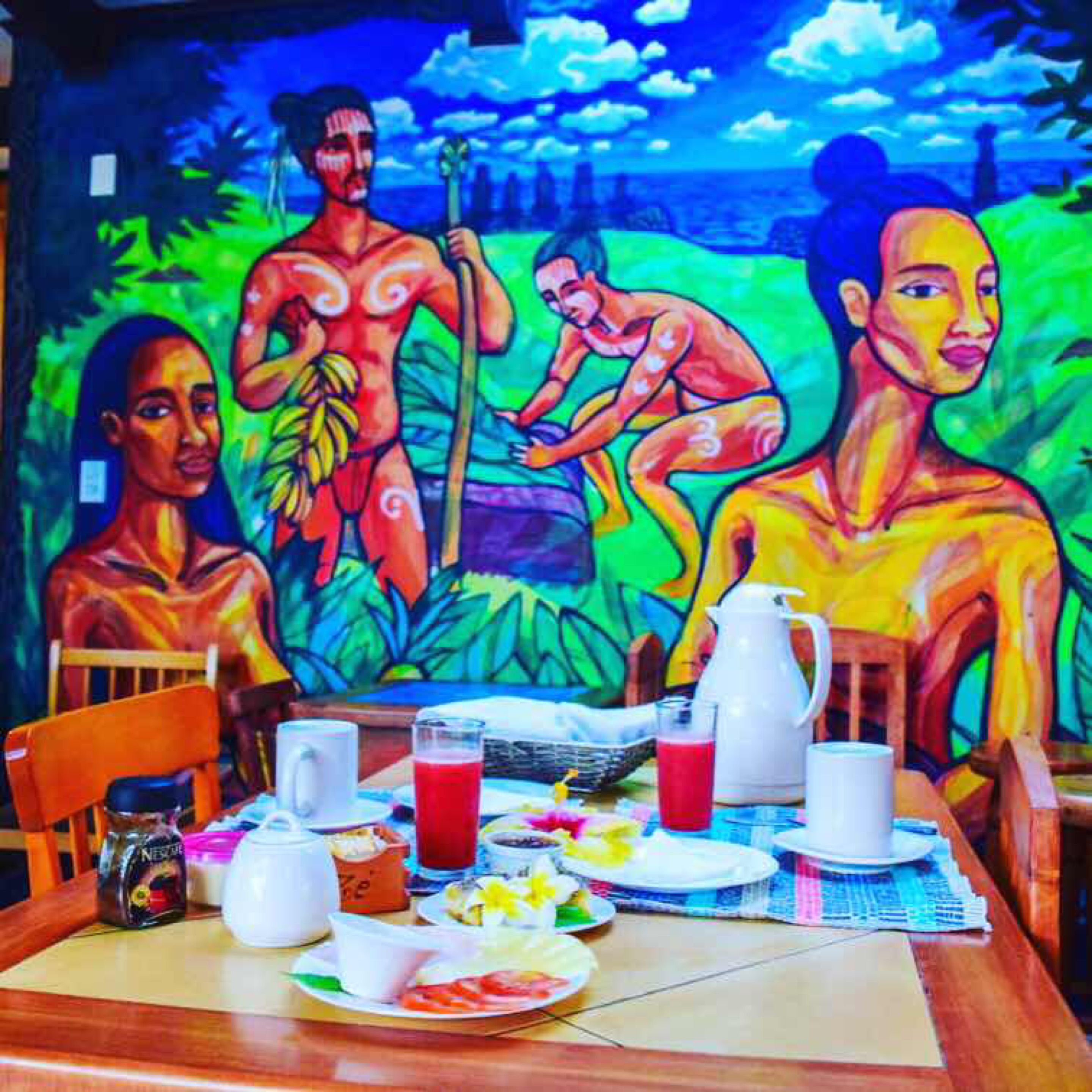 Uma área de refeições com um mural de figuras tropicais, uma mesa de madeira com pratos, copos e um jarro de suco.