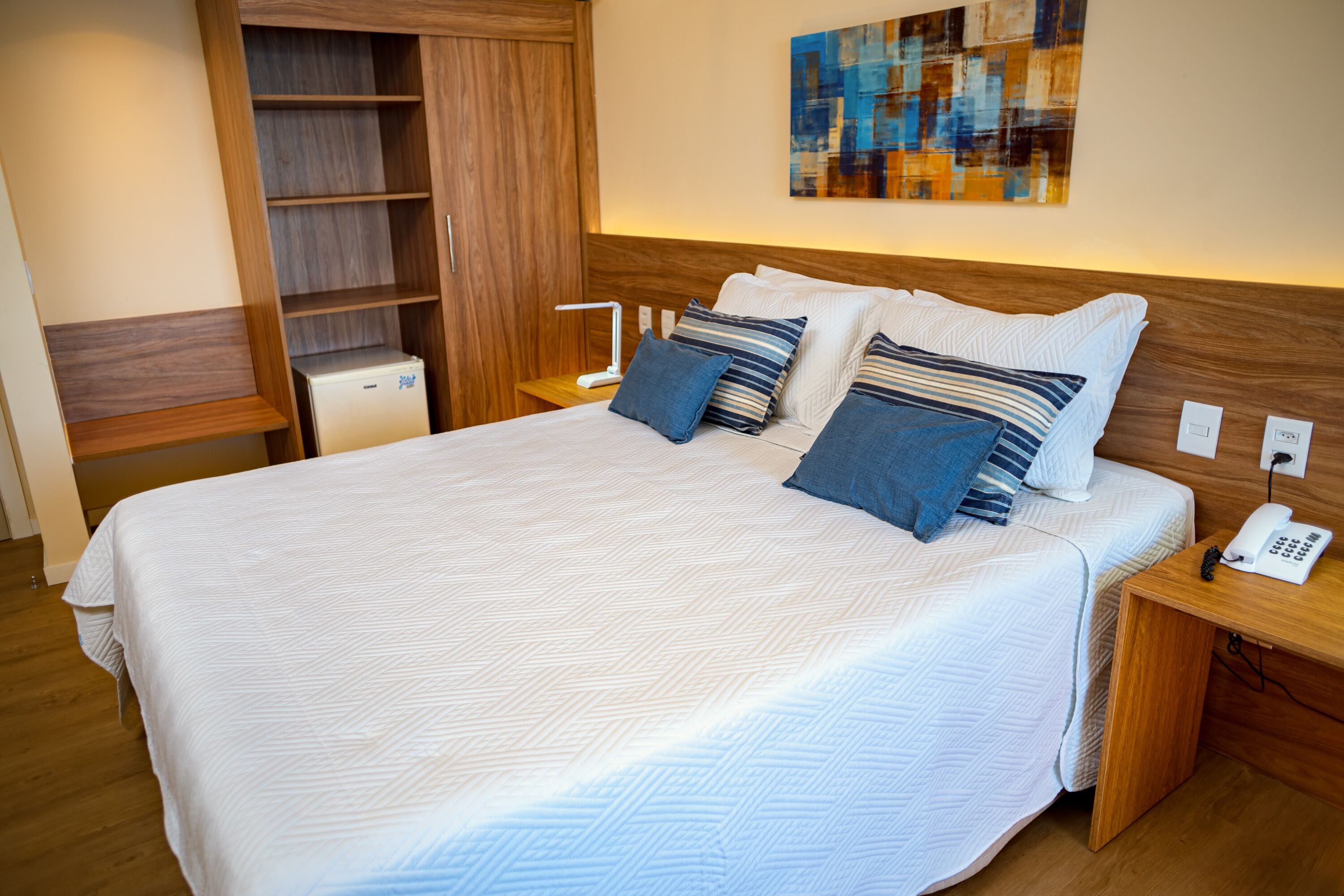 A neatly made bed with a white and blue striped pillow set, a bedside table with a lamp, and a wooden wardrobe.