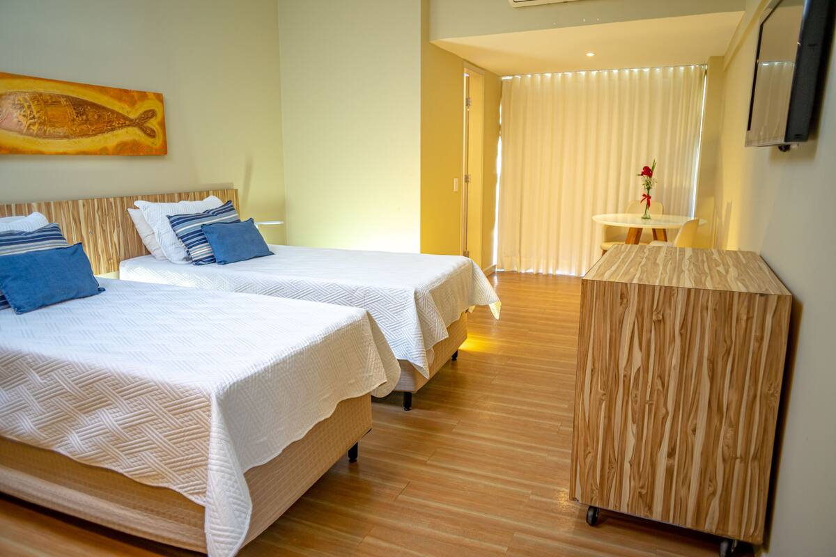 Een hotelkamer met twee bedden, een houten vloer, een tafeltje met een vaas en een televisie aan de muur.