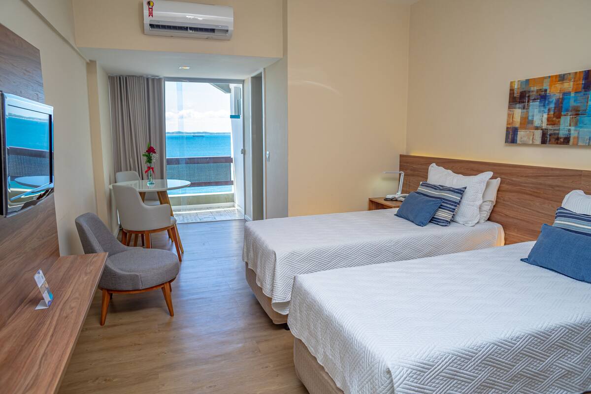 Een hotelkamer met twee bedden, een bureau, een stoel, een televisie en een balkon met uitzicht op zee.