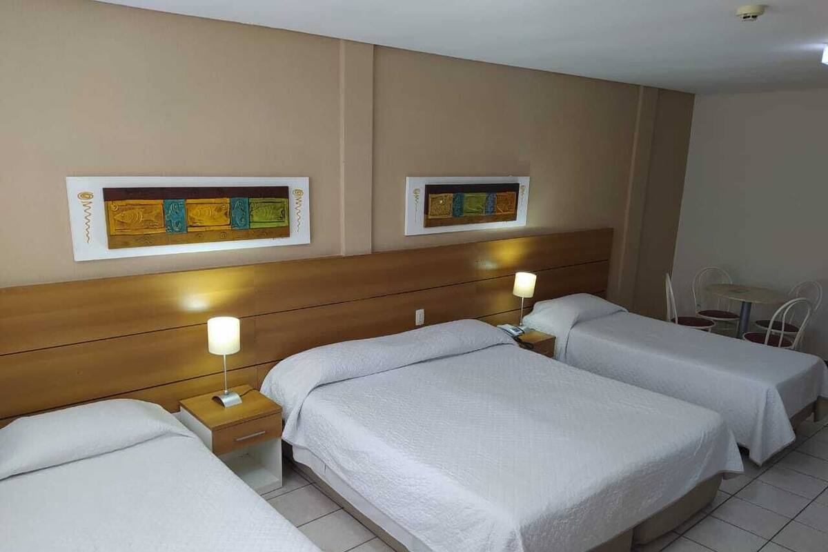 Een hotelkamer met drie bedden, een houten hoofdeinde, een klein tafeltje met een stoel en twee kunstwerken aan de muur.