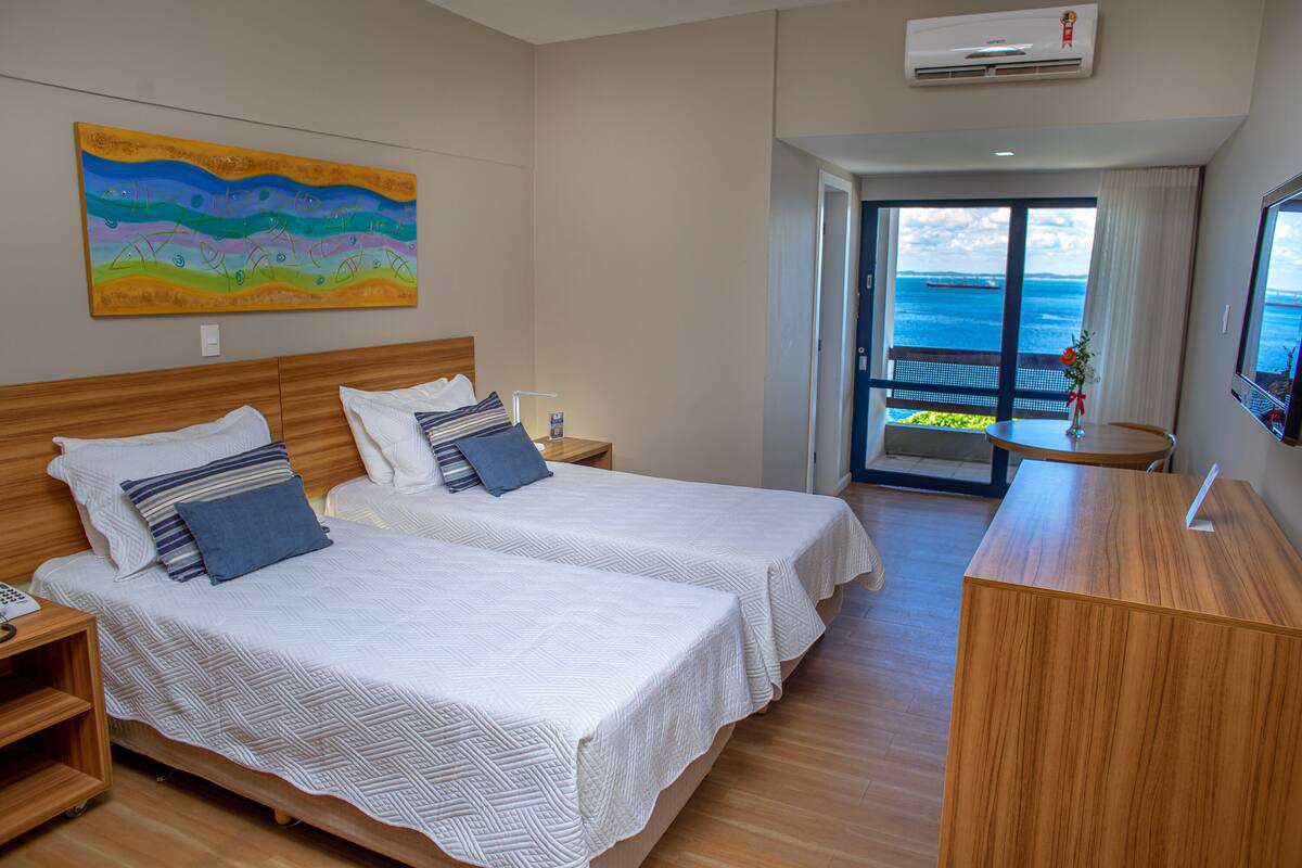 Een hotelkamer met twee bedden, een houten hoofdeinde, een tafeltje en uitzicht op zee door een glazen deur.