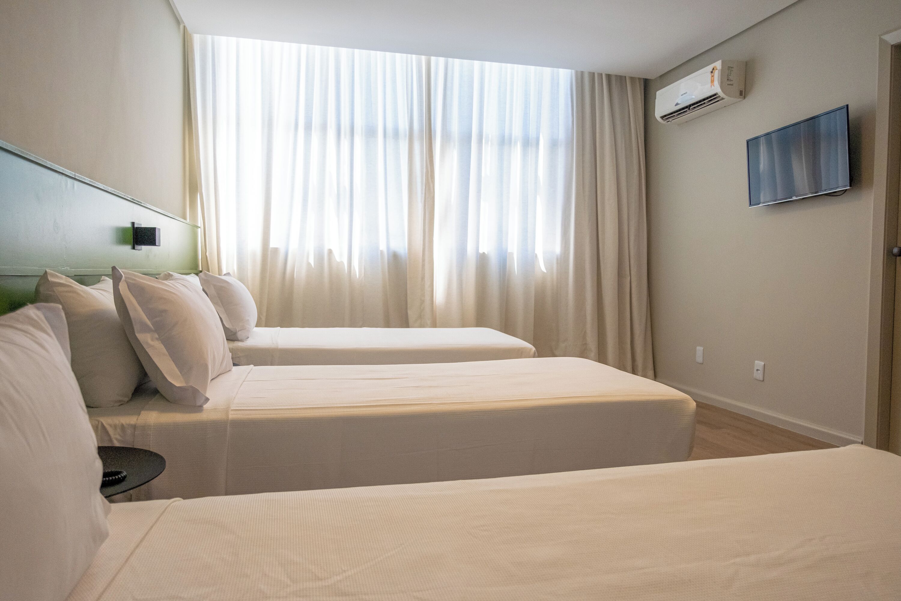 A hotel room with two beds, a flat-screen TV, an air conditioning unit, and a large window with curtains.