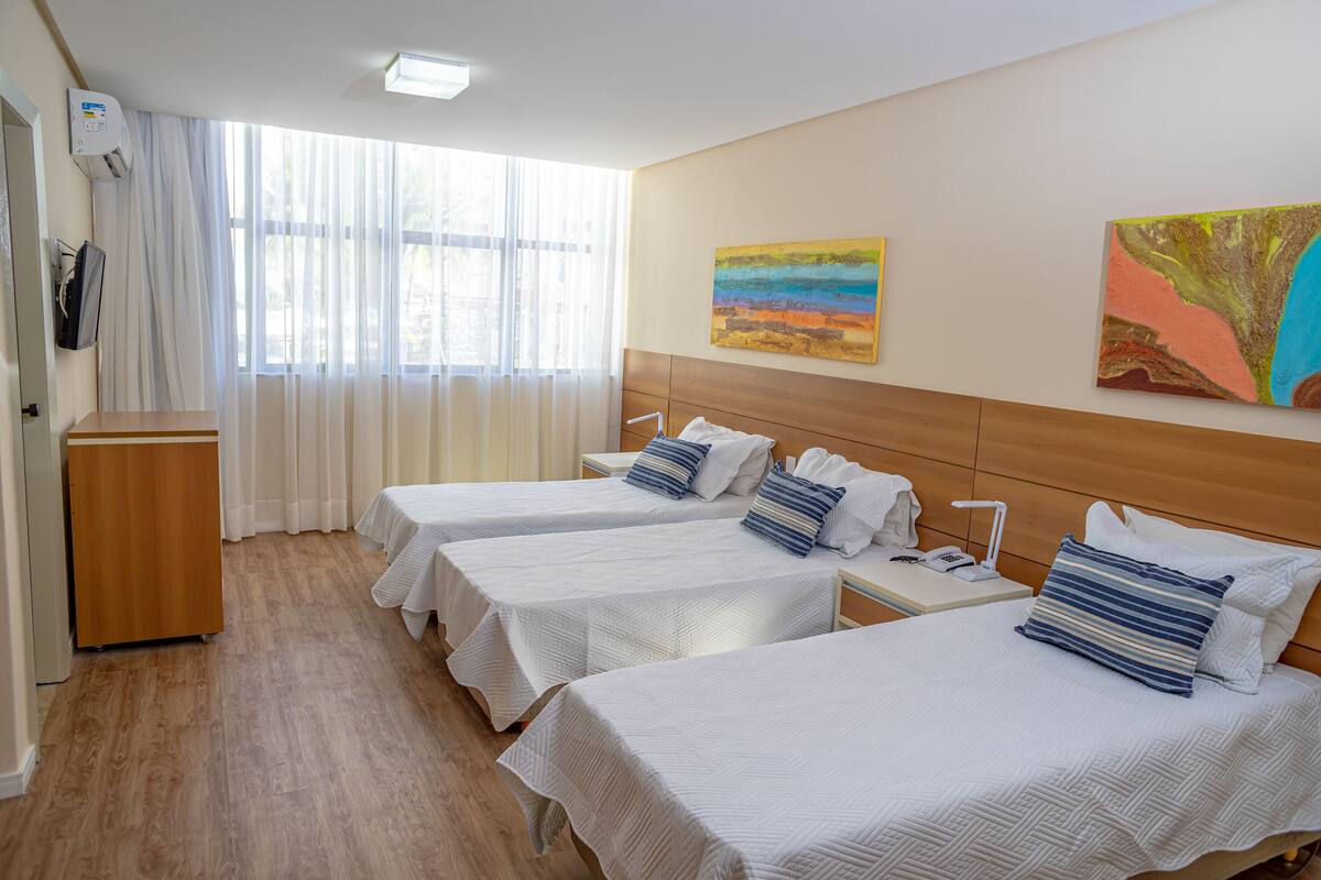 Een hotelkamer met drie eenpersoonsbedden, een houten hoofdbord, een nachtkastje met een lamp en een groot raam met dunne gordijnen.