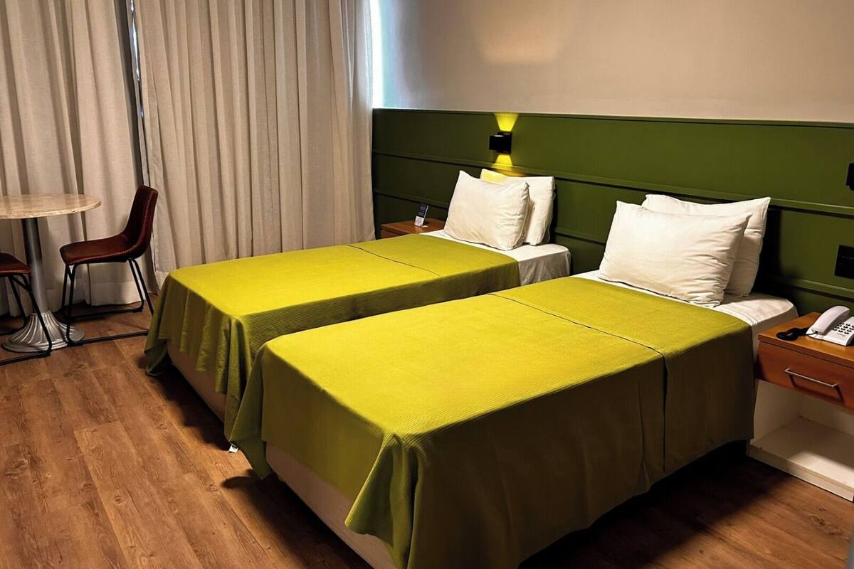 Een hotelkamer met twee eenpersoonsbedden, een tafeltje met een stoel, een nachtkastje met een telefoon en een raam met gordijnen.