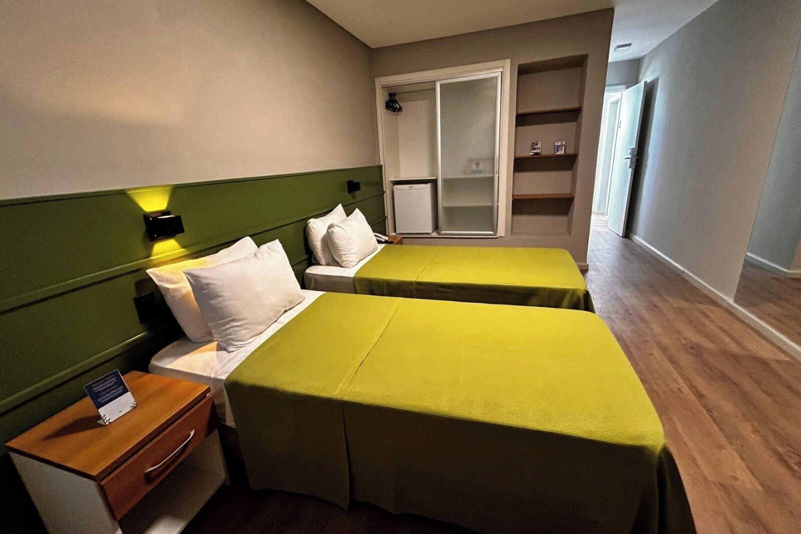 Habitación de hotel con dos camas individuales, una mesita de noche de madera, un escritorio pequeño y un armario con espejo.
