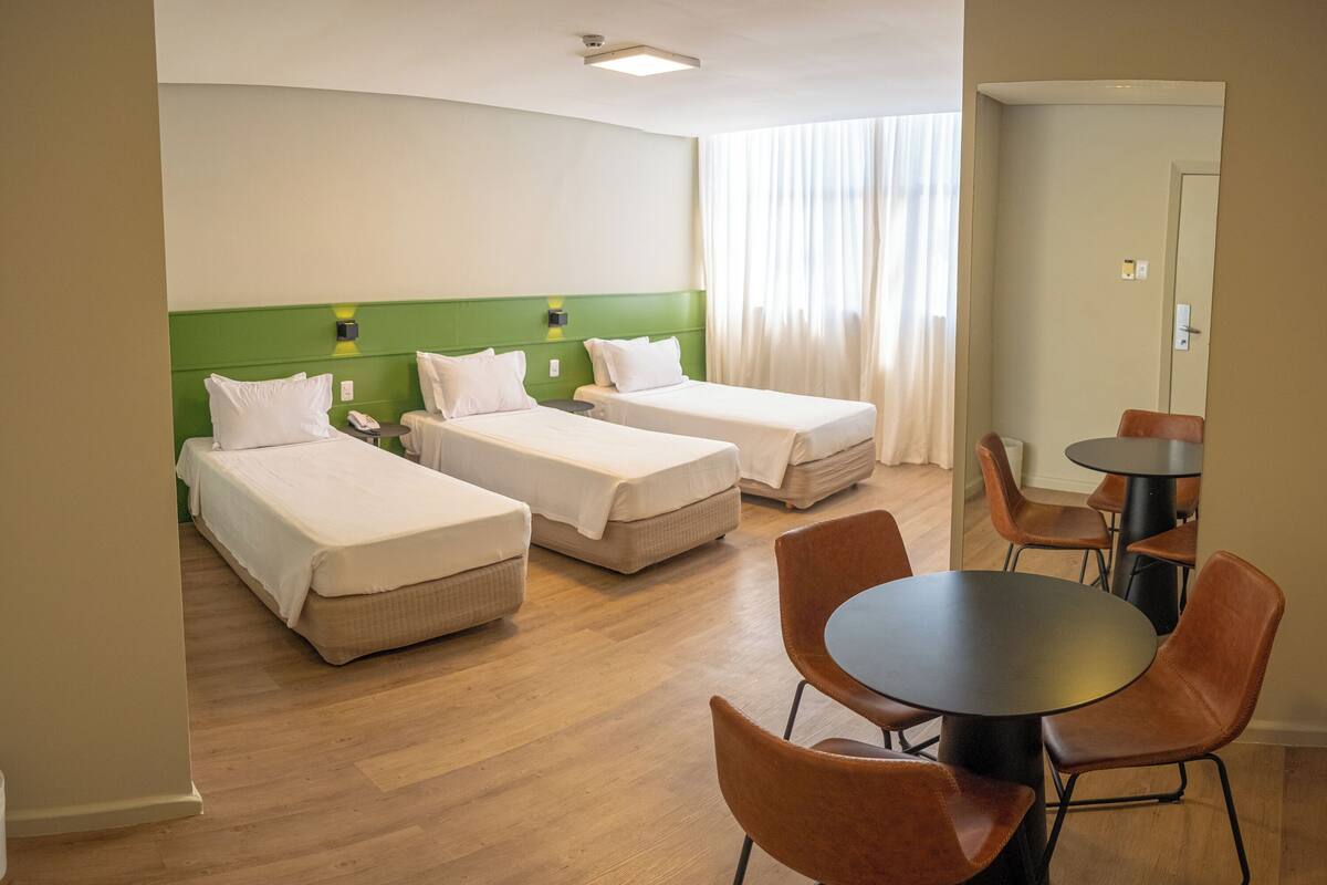 Een hotelkamer met drie enkele bedden, een eettafel met stoelen en een groot raam met gordijnen.