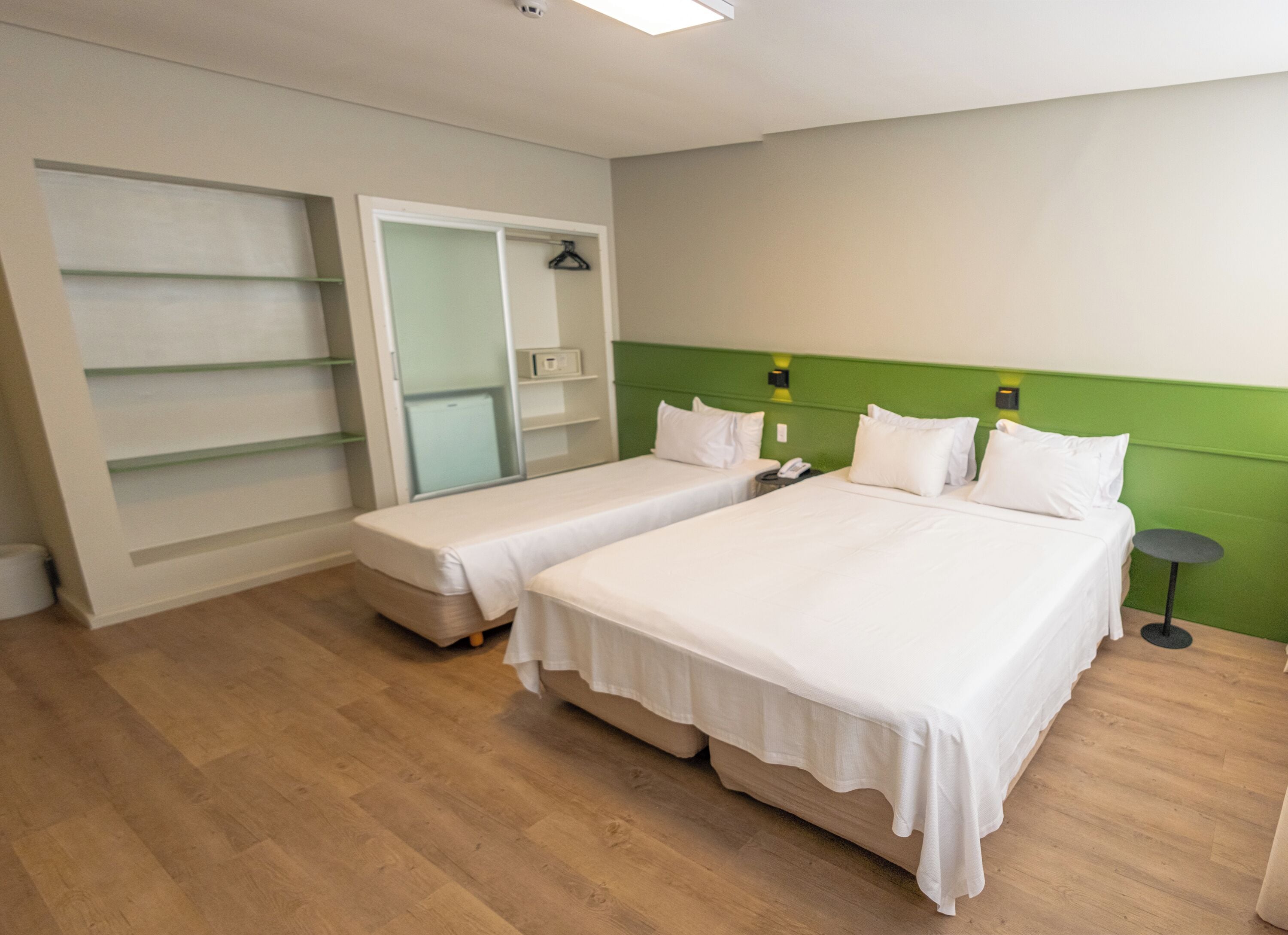 Una habitació d'hotel amb dos llits individuals, una paret d'accent verd, una petita taula de nit i un armari amb portes de vidre.