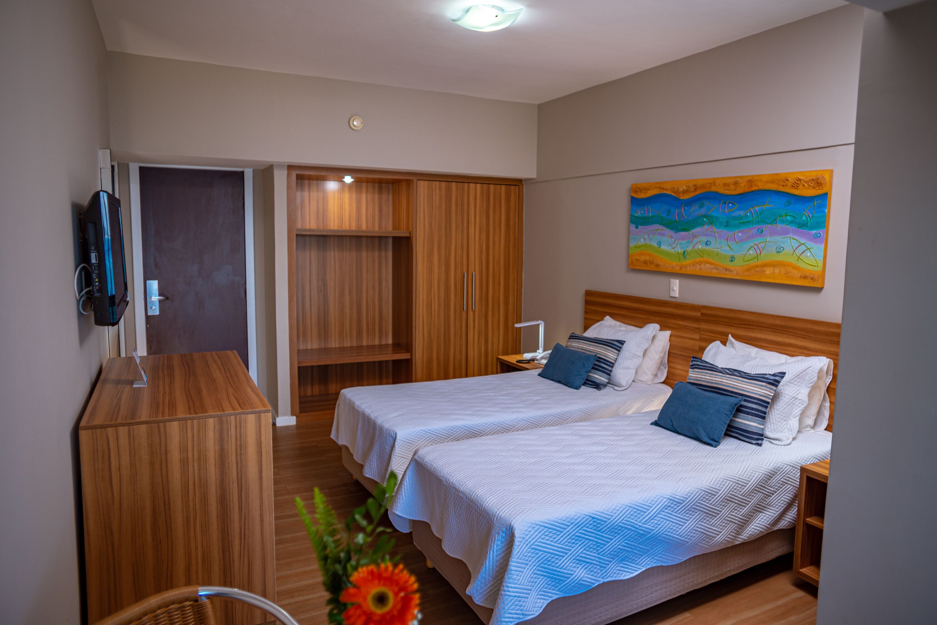 A hotel room with two beds, a wooden wardrobe, a desk, and a painting on the wall.
