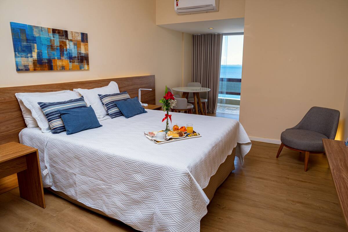 Een hotelkamer met een groot bed, een eettafel, een stoel en uitzicht op zee.