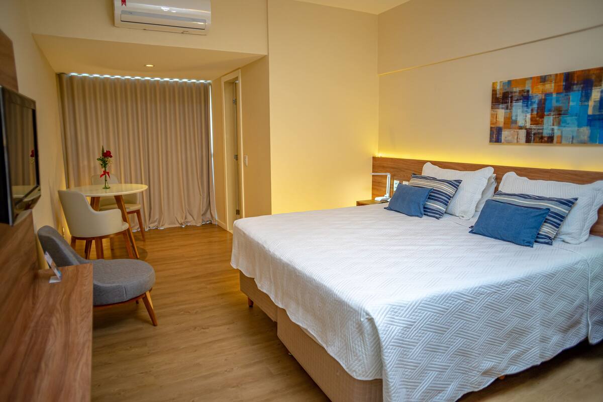 Een hotelkamer met een groot bed, een bureau met stoel, een klein tafeltje met vaas en een gordijn.
