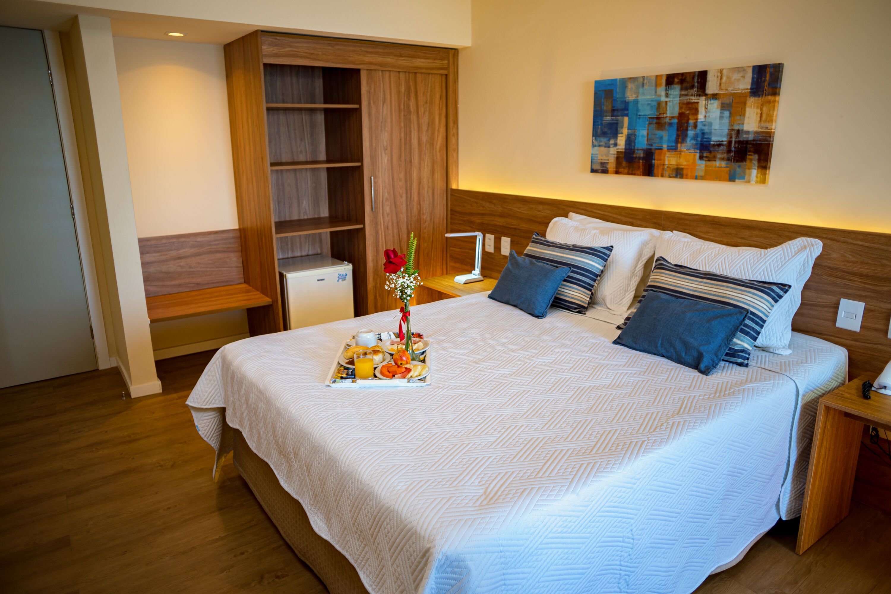 A neatly made bed with a tray of breakfast items, a wooden wardrobe, and a modern abstract painting on the wall.