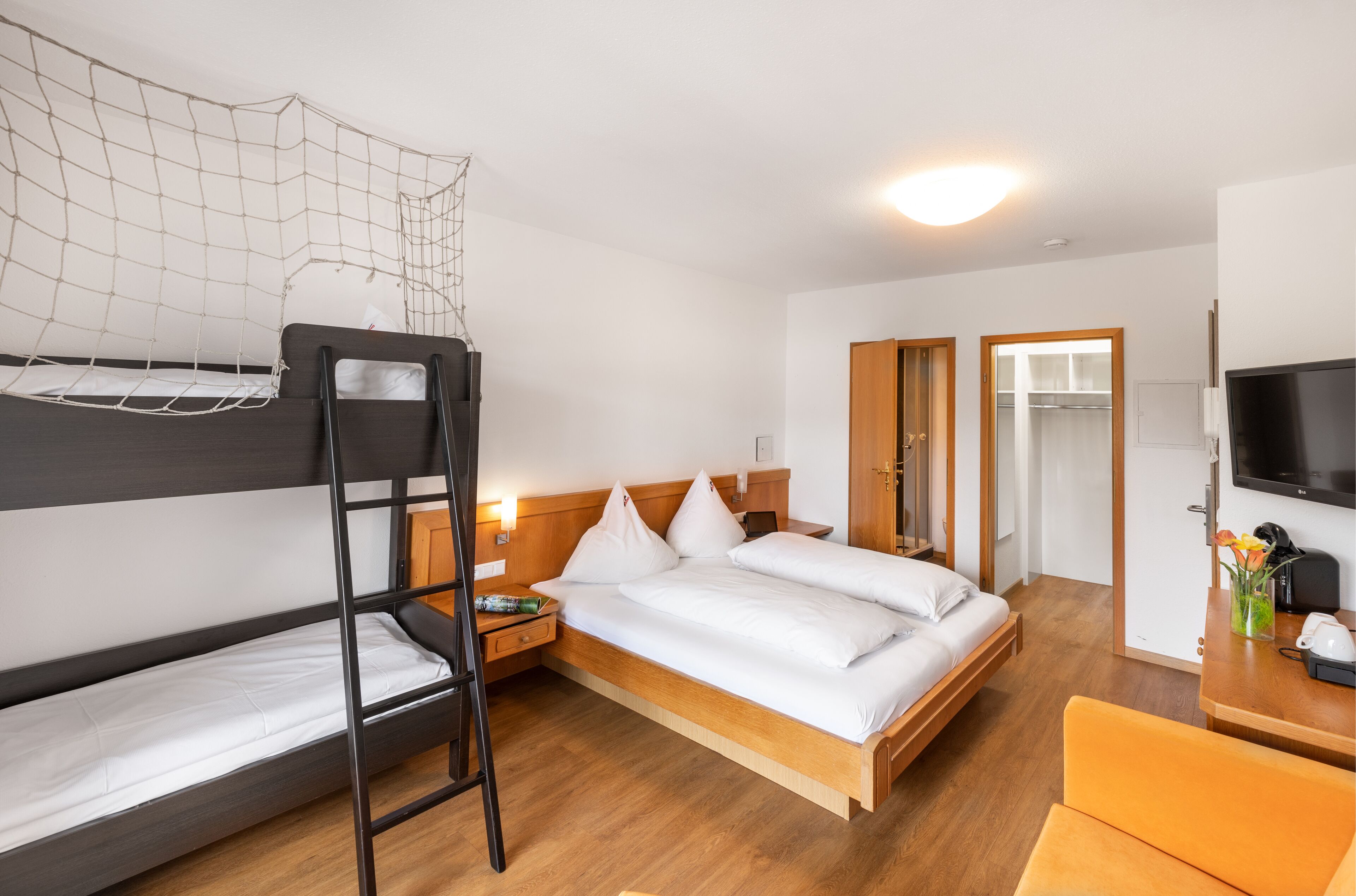 Ein Zimmer mit einem Etagenbett, einem Einzelbett, einem Schreibtisch mit Stuhl, einem Fernseher und einem durch eine offene Tür einsehbaren Badezimmer.