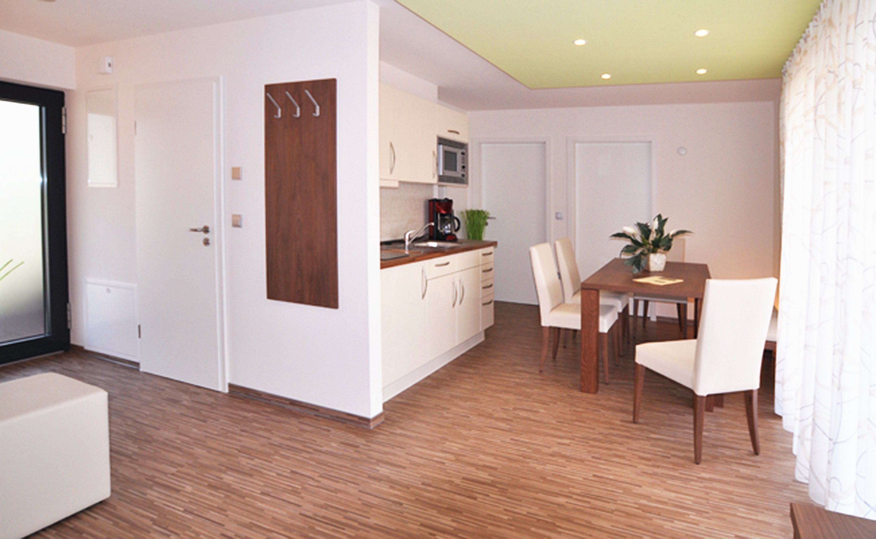 A modern kitchen with white cabinets, a wooden dining table, and chairs. There is a microwave, a coffee maker, and a potted plant on the counter.