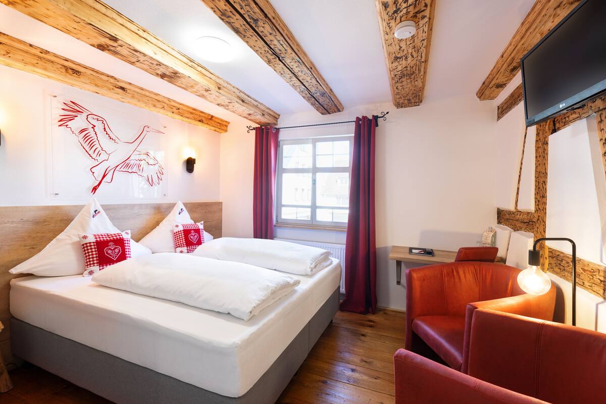 A hotel room with a wooden ceiling, a bed with white linens, red checkered pillows, a red chair, and a television mounted on the wall.