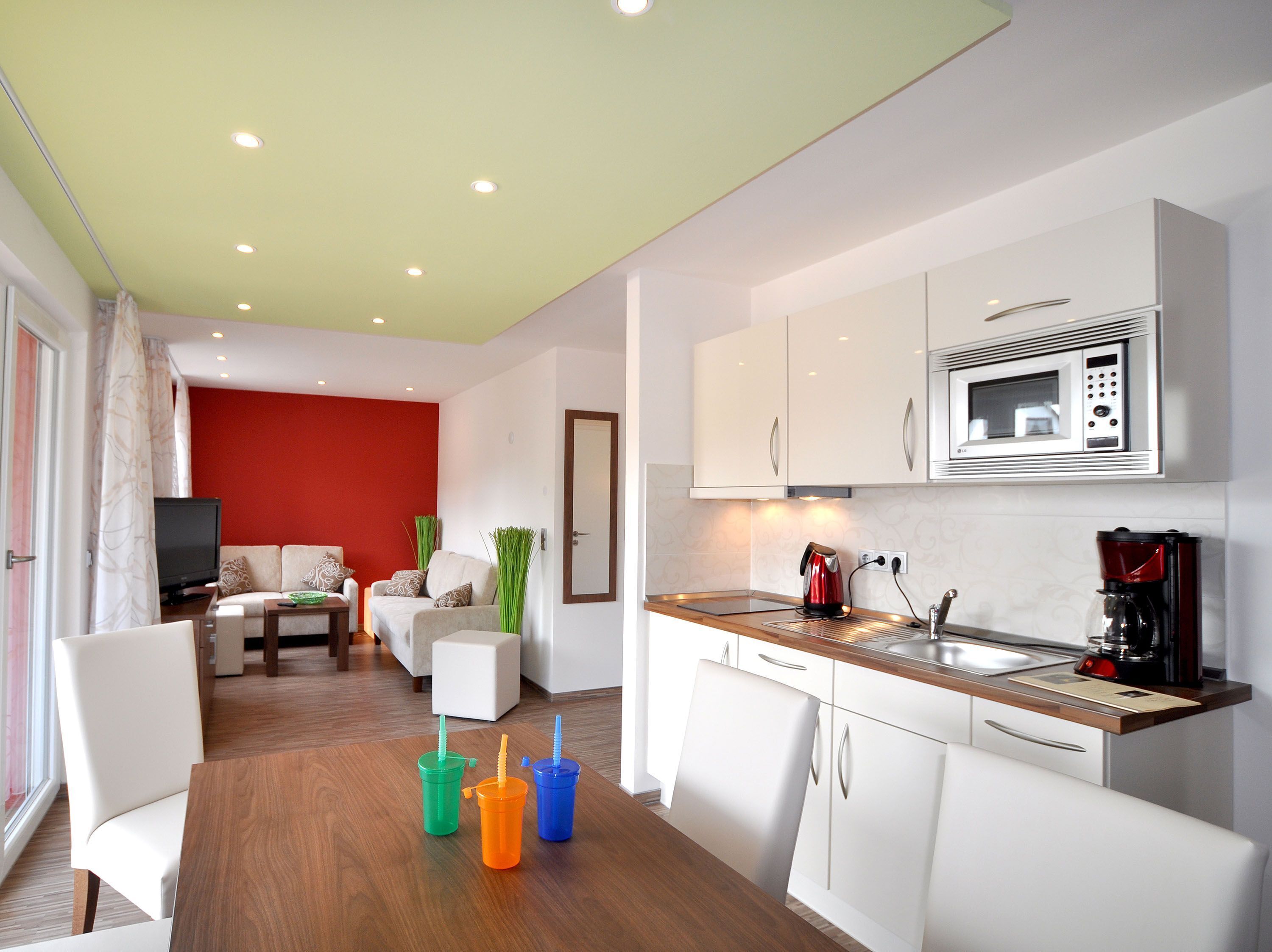 A modern kitchen with white cabinets, a microwave, and a coffee maker. There is a dining area with a table and chairs, and a living room with a sofa and a television.