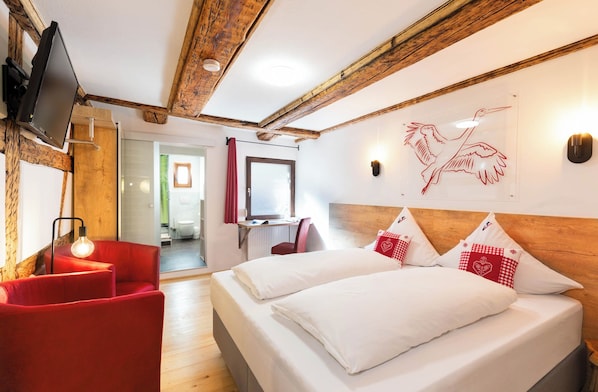 Habitación de hotel con techo de madera, cama con ropa blanca y almohadas rojas, un sillón rojo, un escritorio con una silla y un televisor montado en la pared.