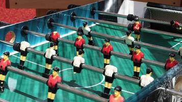 Una mesa de fútbol de mesa con jugadores con uniformes rojos y blancos, una mesa de billar al fondo, y una vista parcial de una habitación con alfombra estampada.