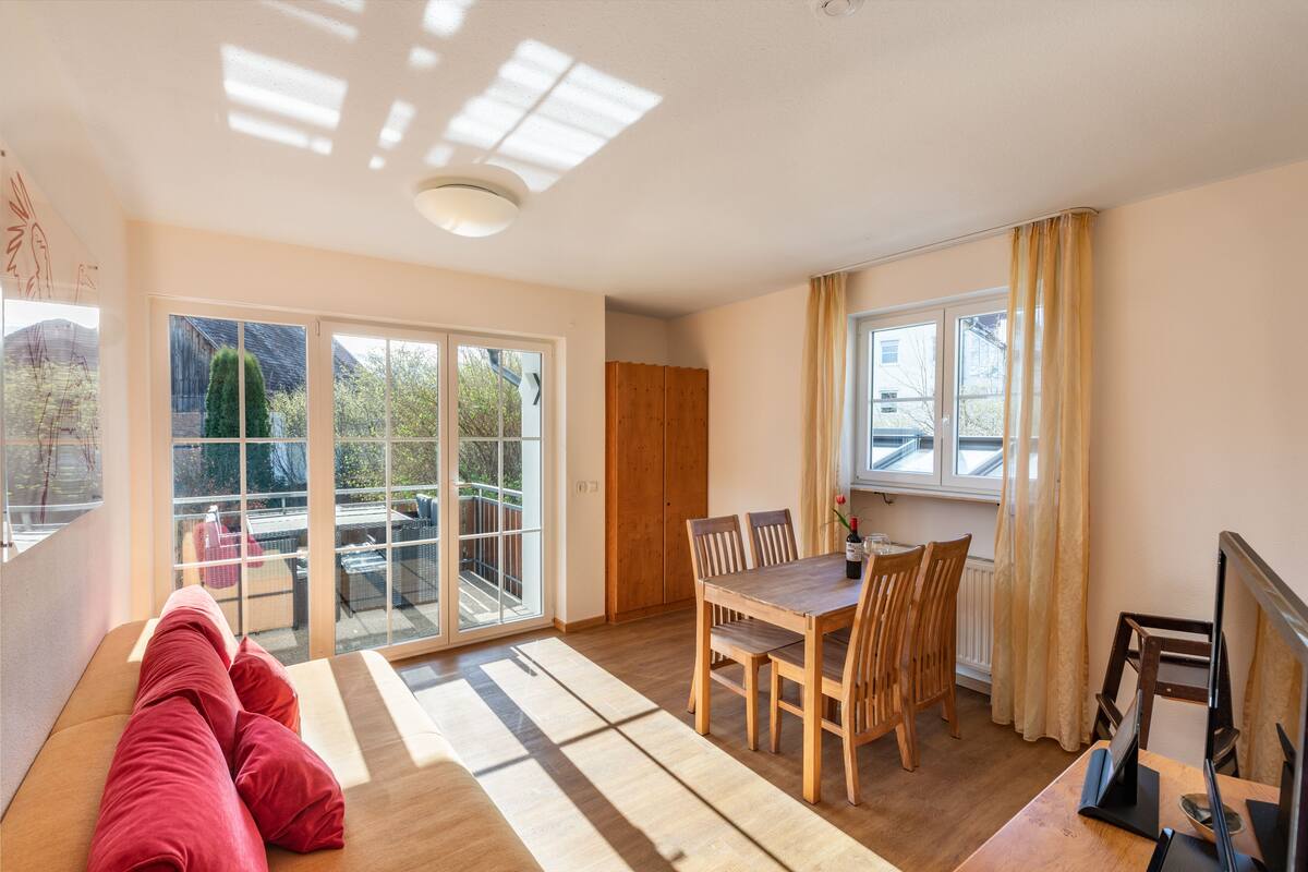 A spacious room with a wooden dining table, chairs, and a sofa. There is a sliding glass door leading to an outdoor area with a view of trees and a building.
