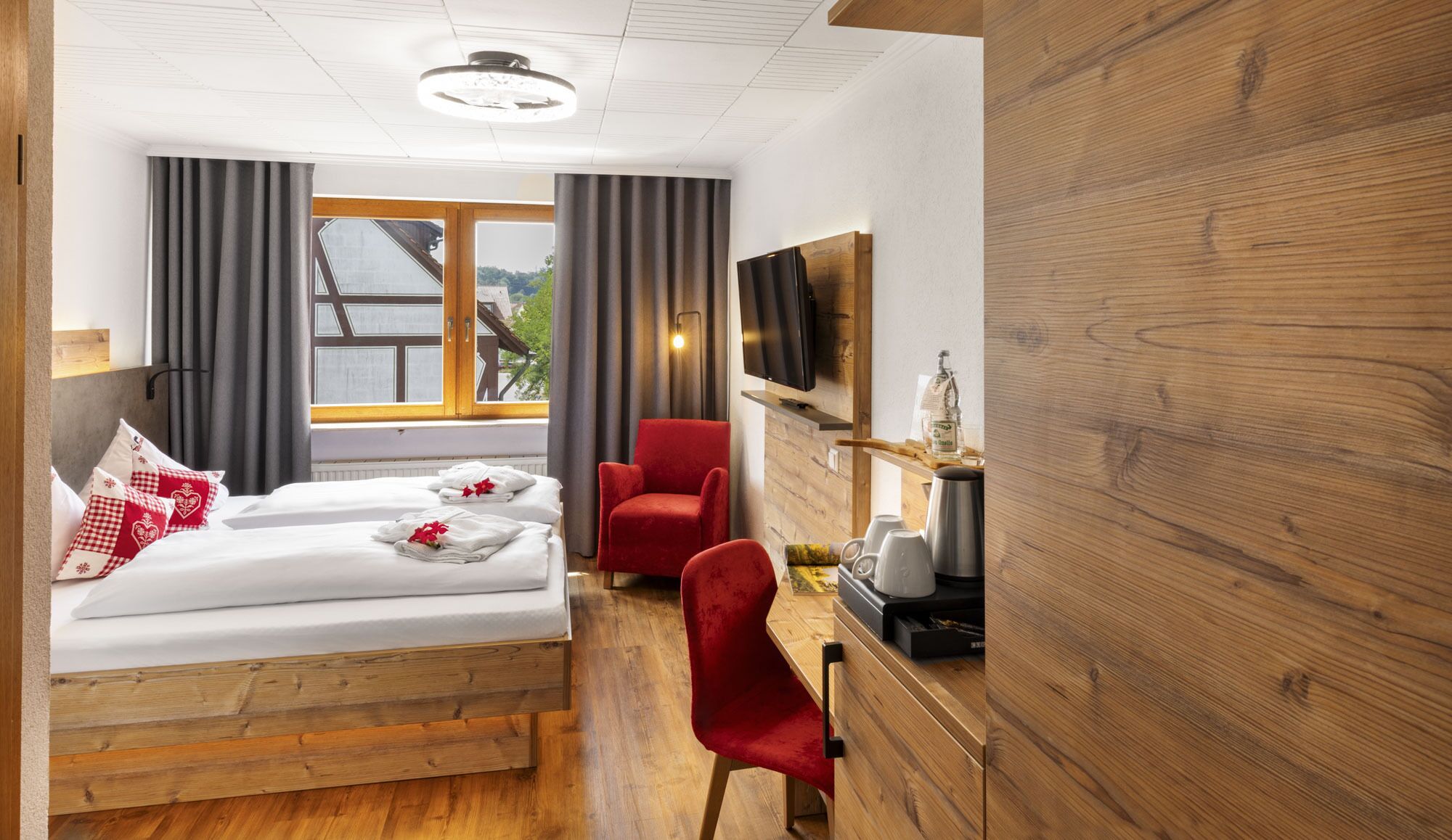 Habitación de hotel con dos camas, una silla roja, un escritorio con cafetera, un televisor y una ventana con cortinas.