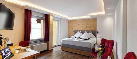 A modern hotel room with a wooden floor, a bed with white bedding, a red armchair, and a desk with a computer and a coffee machine.