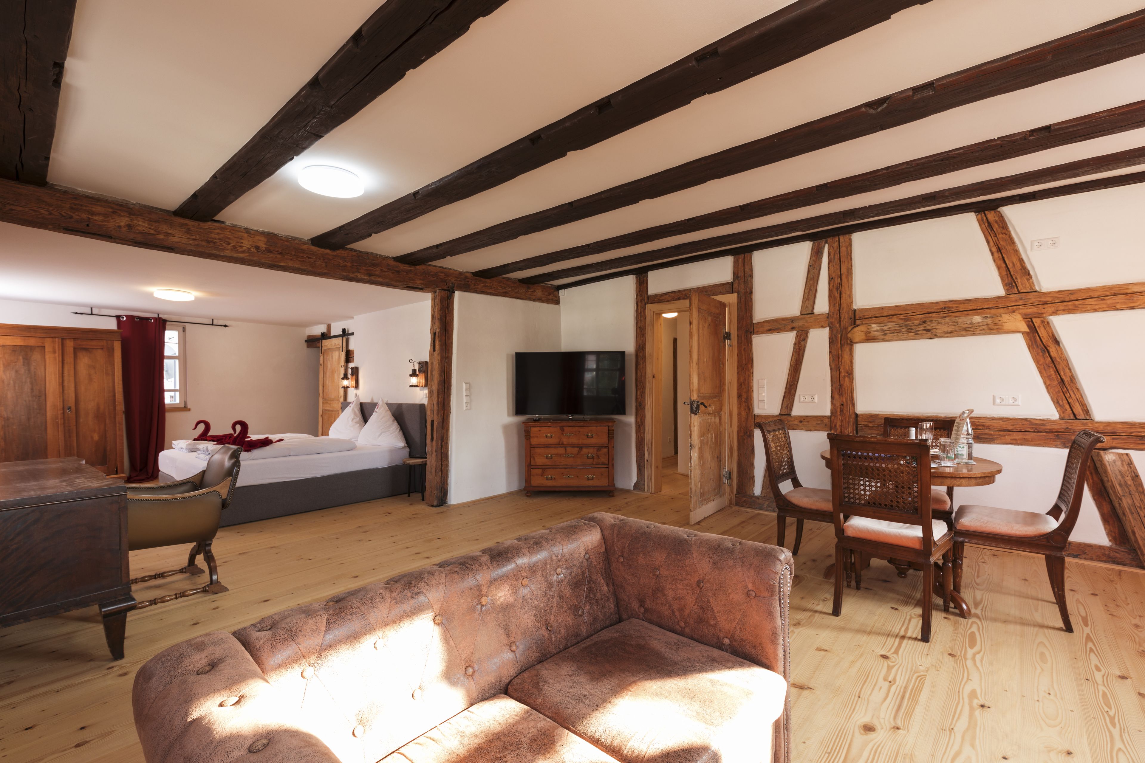 A room with exposed wooden beams, a bed, a television, a dining table with chairs, and a leather sofa.